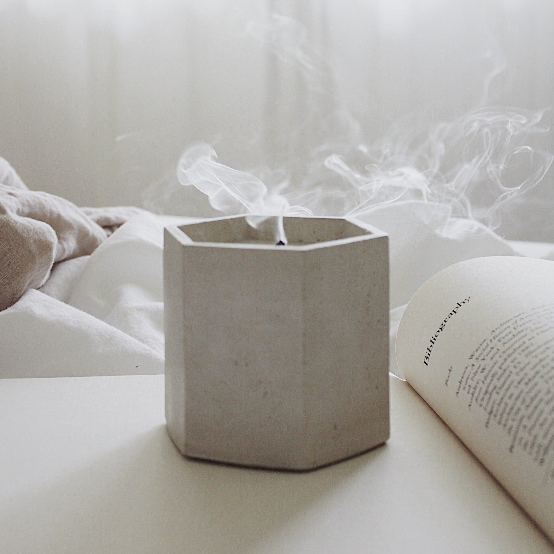 Hexagon white concrete jar with candle sitting on book. Candle wick is smoking. Made by Paige Soy Candles