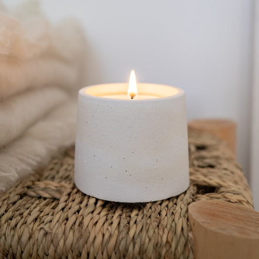 A lit 4 ounce candle on a bench. Jar holding candle is circular and hand-made with white cement. Made by Paige Soy Candles